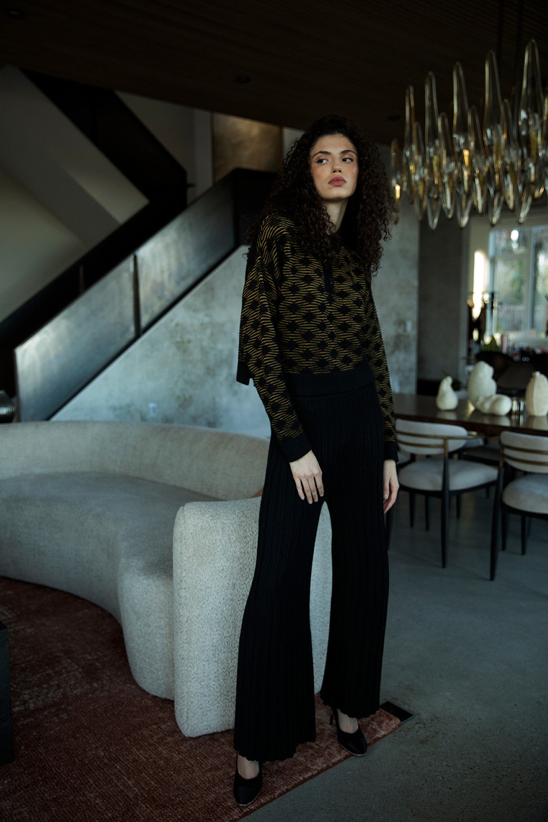 Curly-haired brunette model standing in a living room, wearing the golden black Amelie Polo styled with a French tuck and black alpaca silk stretch trousers.