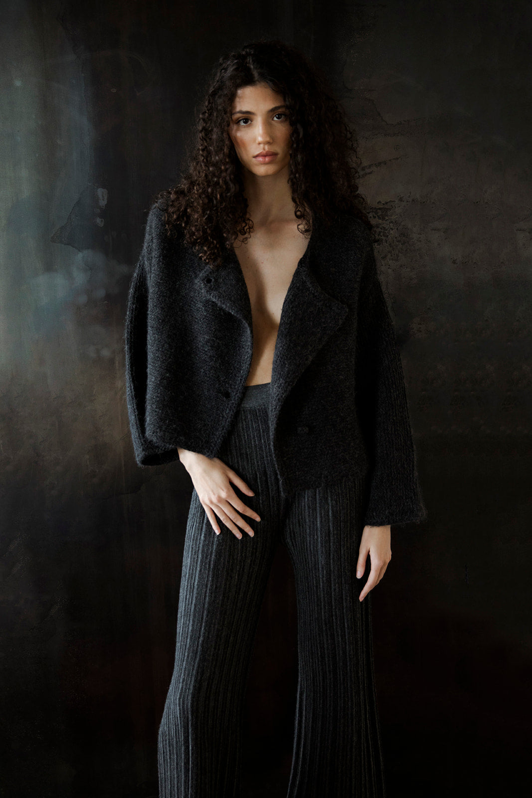 Model standing against a black wall, wearing the Momo Handknit Cardigan in Charcoal open over matching grey trousers known as the Marcelle Trousers by Sskein. Sustainably made in Peru.