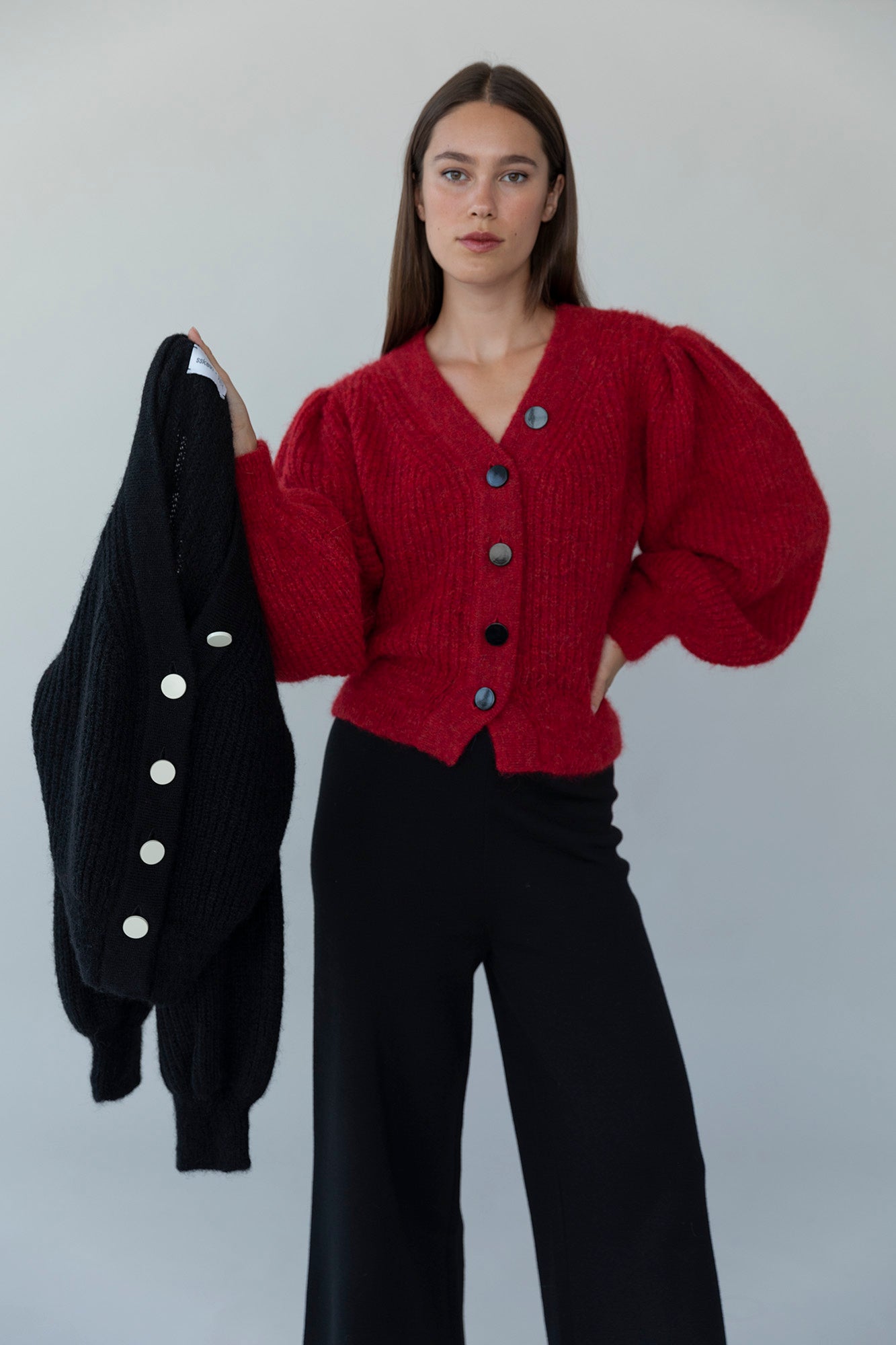 Model wearing the MP Cardigan in crimson red holding out the black version of the cardigan to showcase both color options. Both feature a feminine, dramatic silhouette and are crafted from baby alpaca wool.