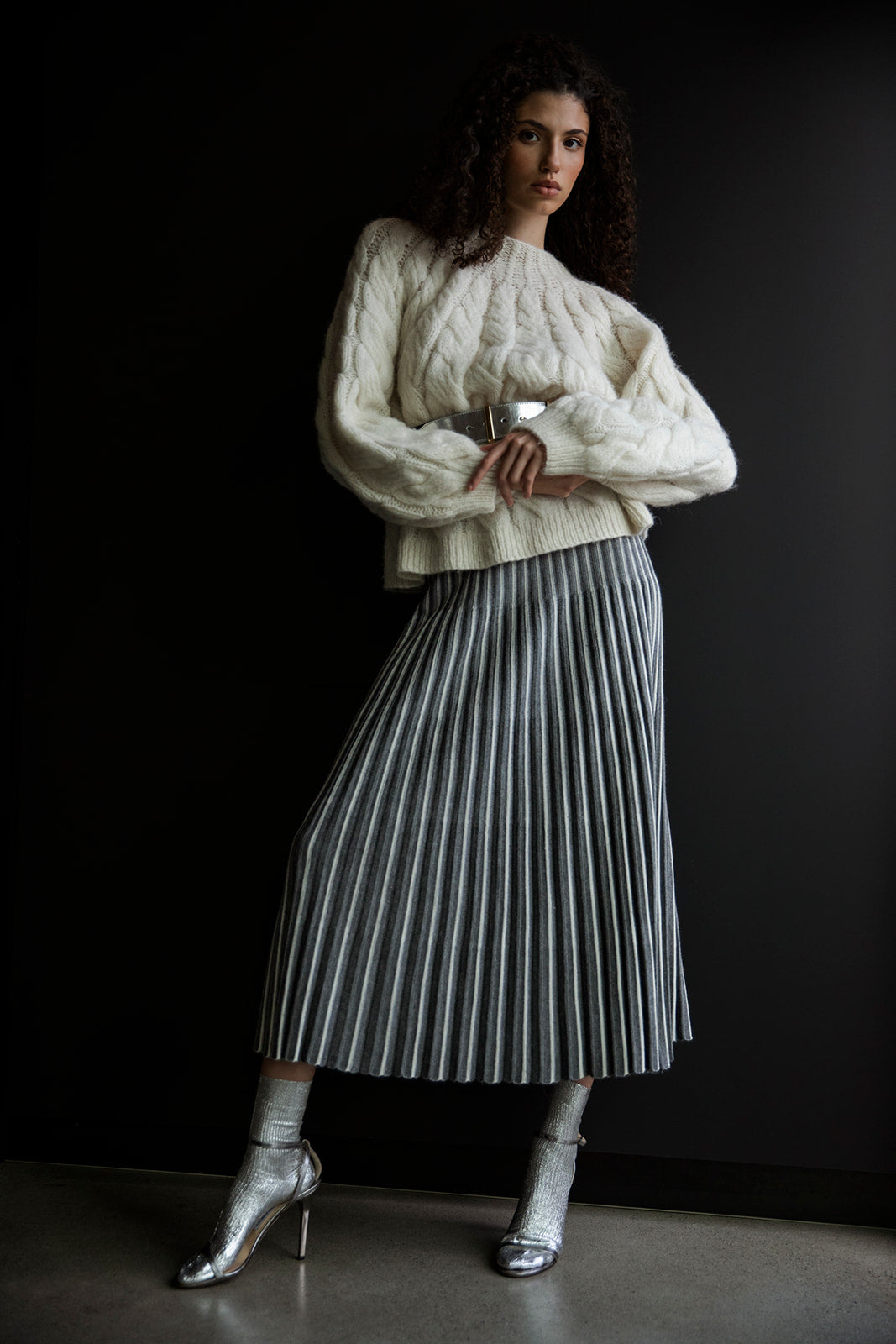 Model confidently crossing her arms in the Adeline Skirt, styled with an ivory handknit sweater by Sskein cinched with a chic silver belt. Complete the look with elegant silver socks and metallic heels for a sophisticated outfit.