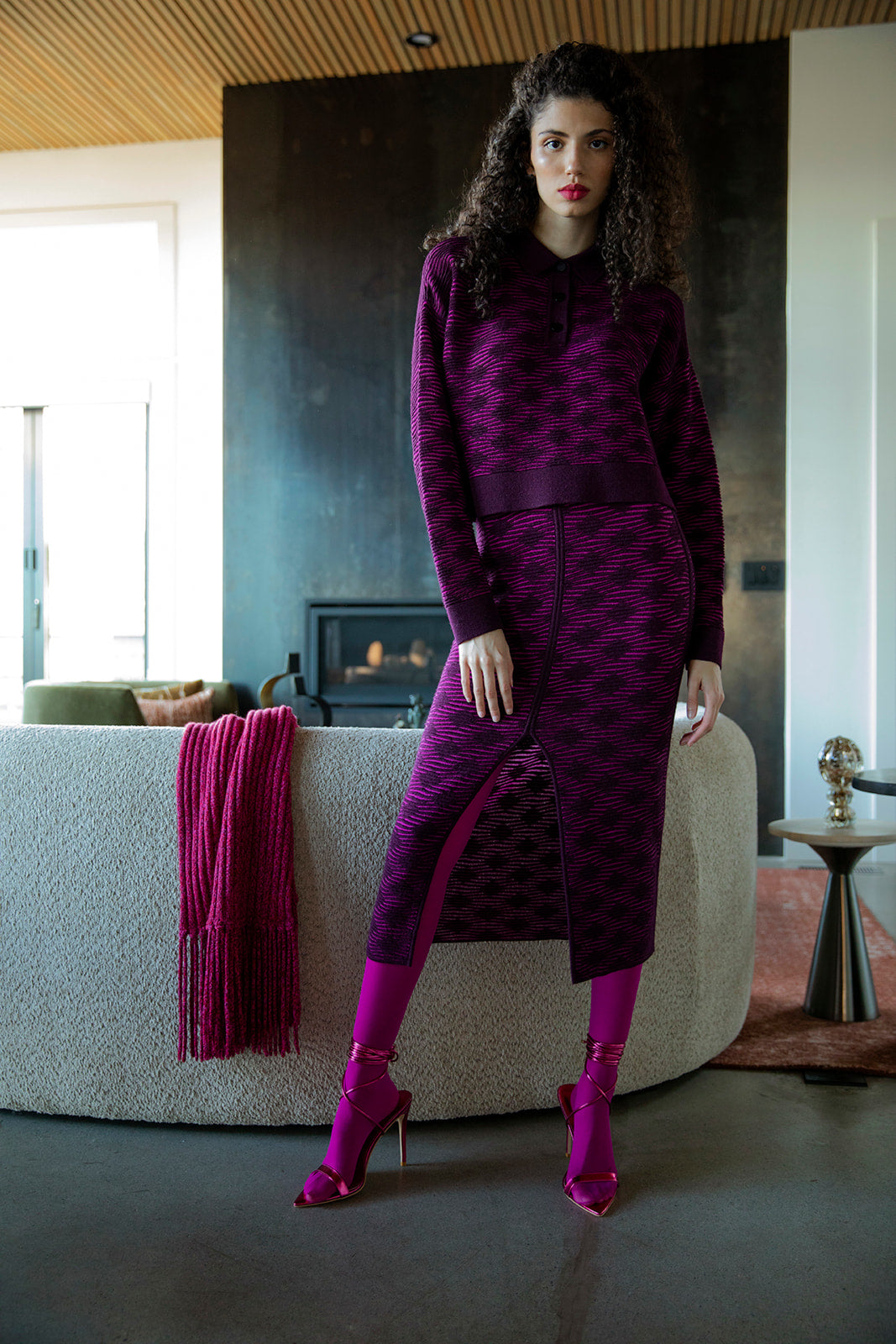 Curly-haired brunette model styled in a complete statement look in fuchsia aubergine. She wears a knitted monochromatic polo sweater and matching pencil skirt. Styled with bright pink tights.