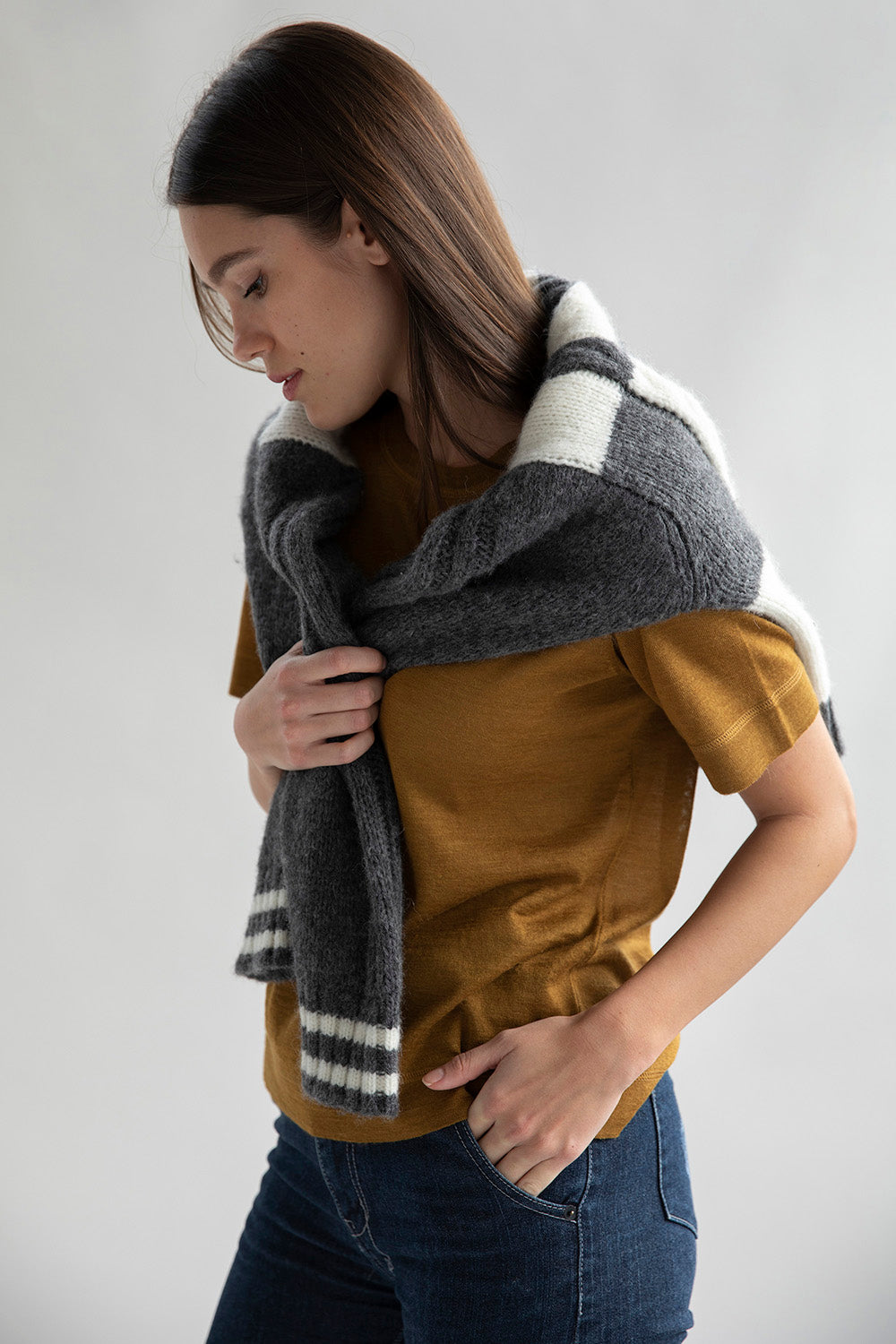 A model looking down wearing a gold sweater tee made from luxury alpaca silk, paired with dark blue denim jeans and a cozy grey plaid sweater wrapped around her shoulders.