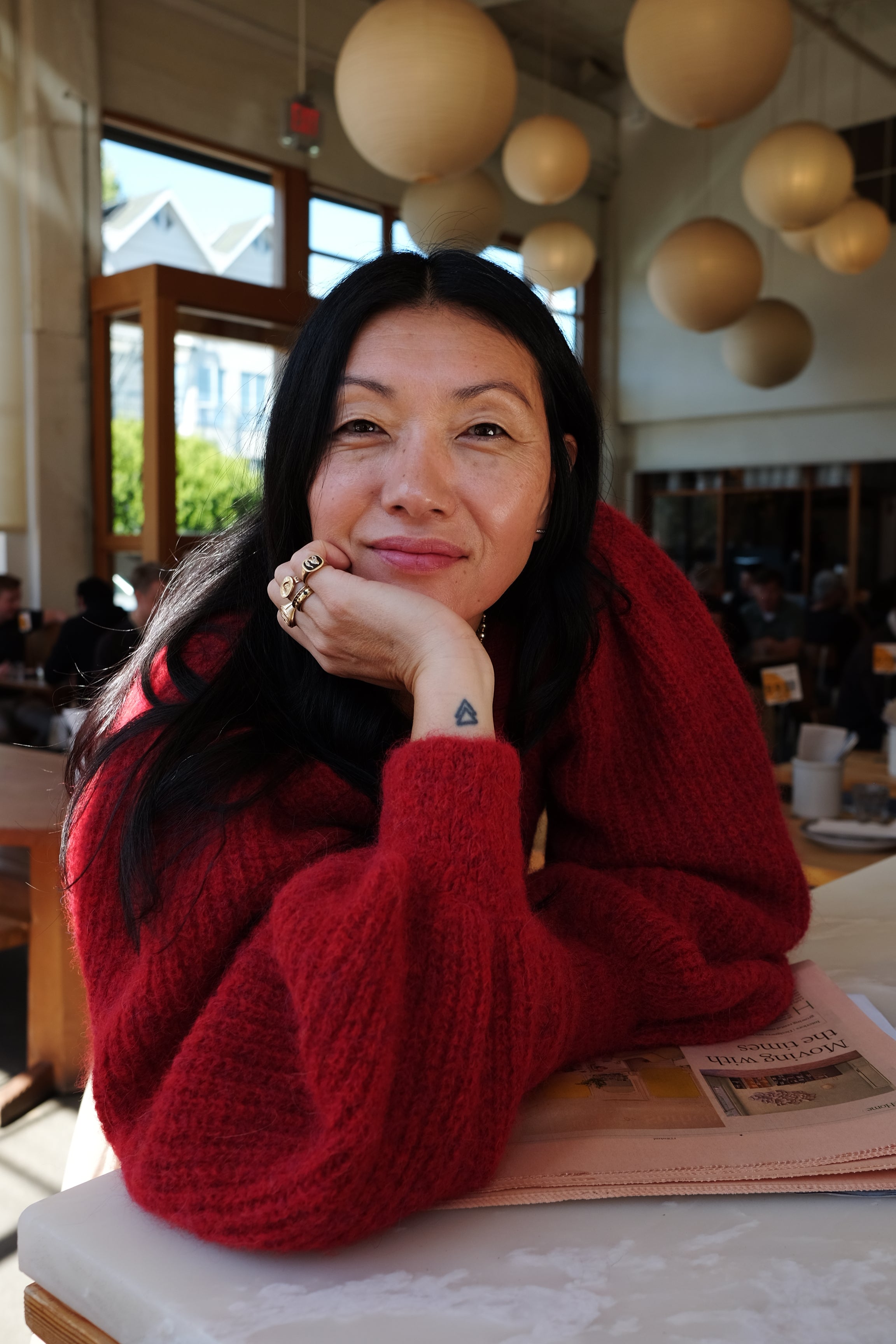 Meredith Peck wearing the sustainably made MP Cardigan in crimson red, smiling with her hand resting under her chin. She is seated at a coffee shop in San Francisco, showcasing the cardigan’s chic and comfortable design.