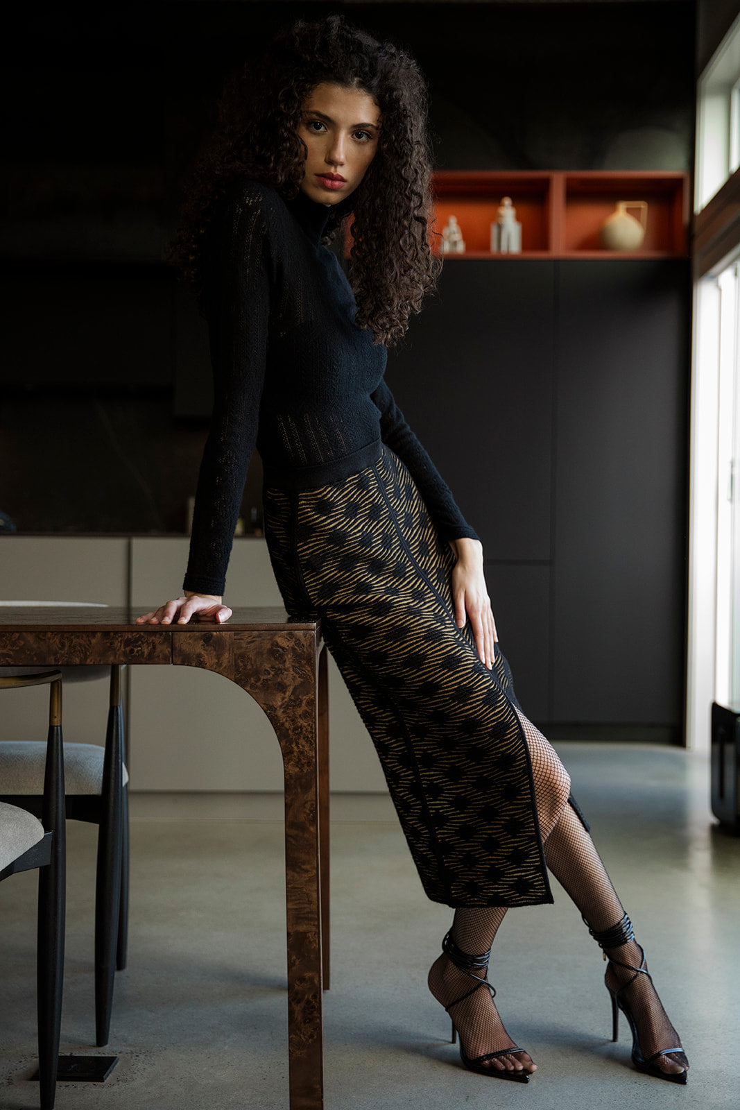 Curly-haired brunette model leaning against a dining table, wearing a golden black pencil skirt paired with a black cashmere turtleneck, fishnet stockings, and black laced-up high heels.