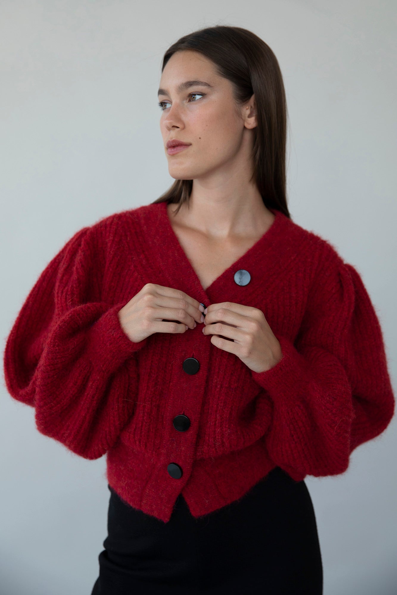 Model wearing the MP Cardigan in crimson red, buttoning up the cardigan’s black buttons. An ethical sweater collaboration between Meredith Peck and Sskein.