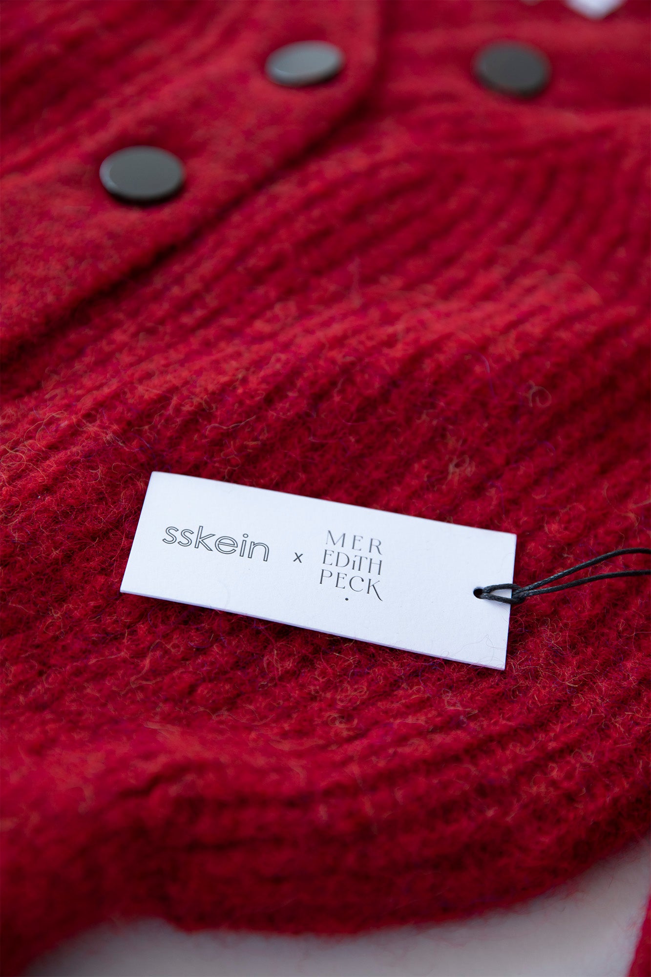 Close-up of the white hang tag featuring the SSKEIN and Meredith Peck logos in black, highlighting the collaboration and sustainable craftsmanship of the MP Cardigan.