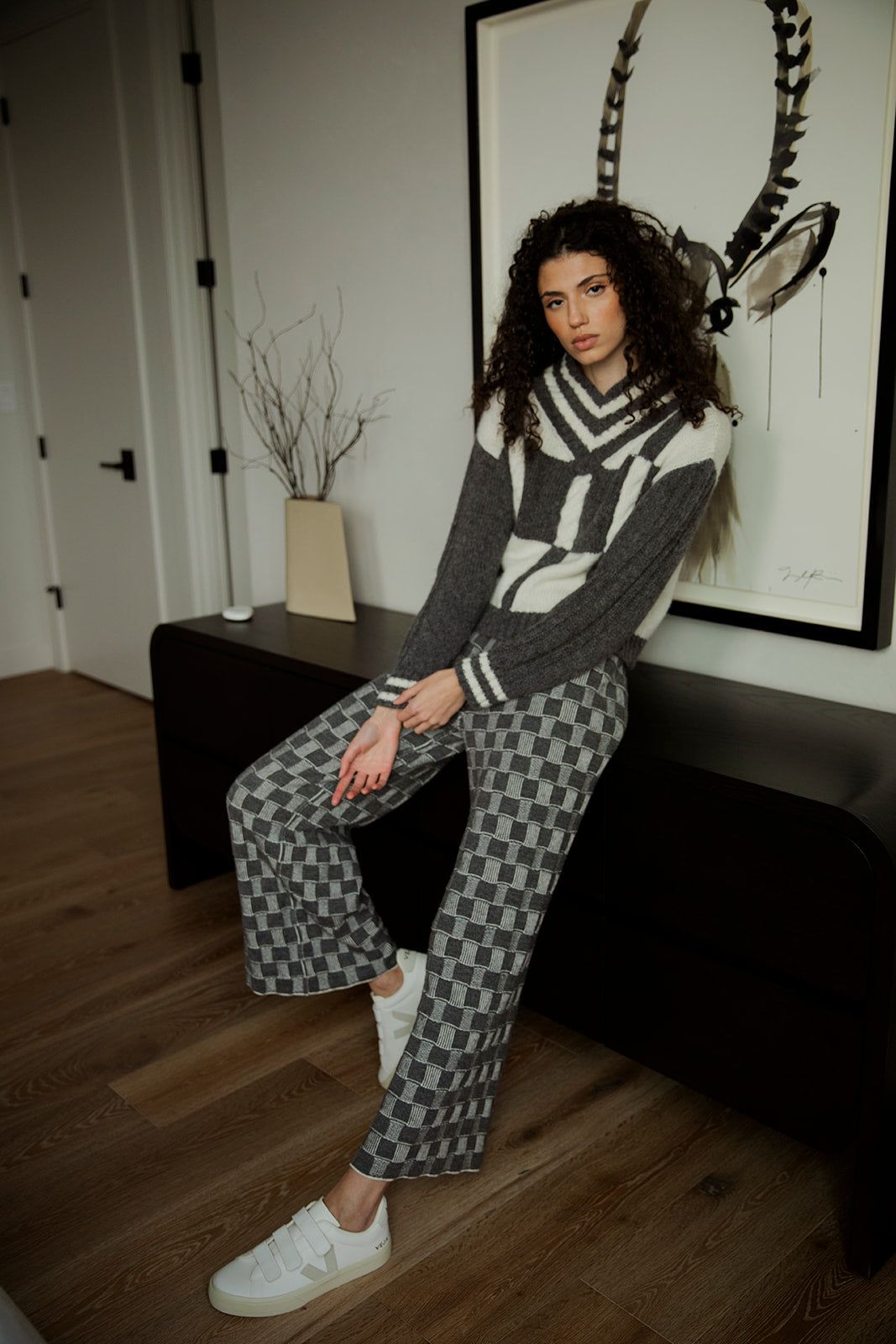 Model styled in Zoe Alpaca Trousers, leaning against a credenza indoors. The ethically made trousers are crafted from 62% baby alpaca and 38% Pima cotton, paired with a grey ivory V Neck Pullover.